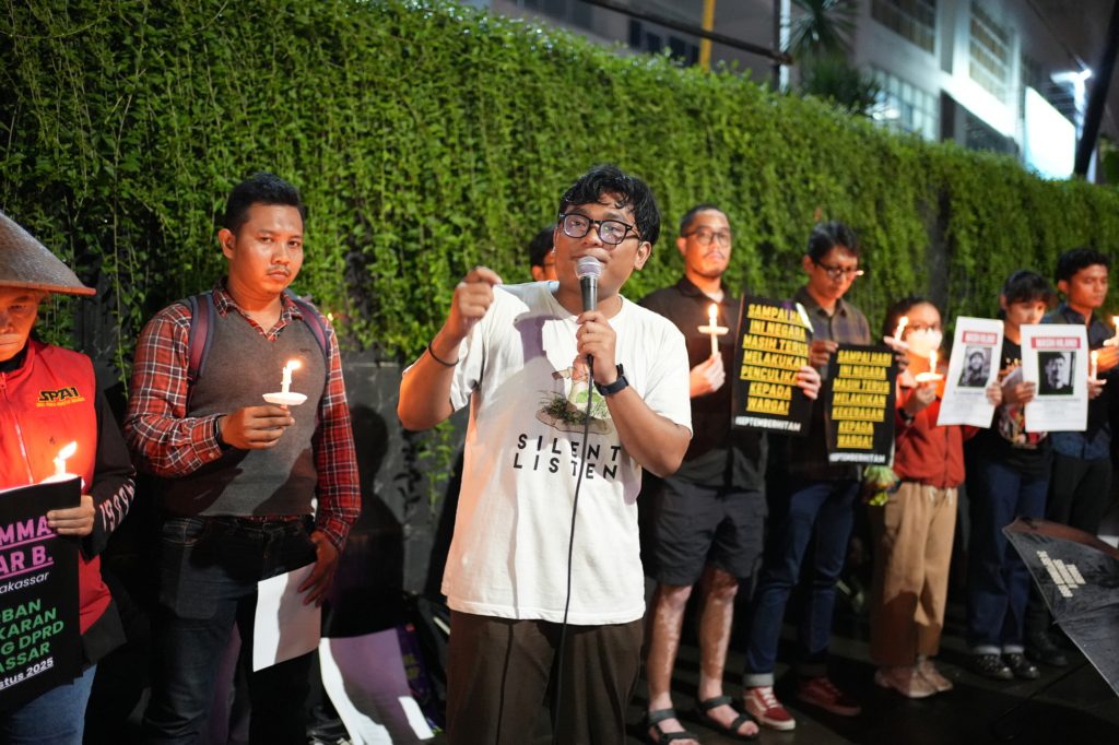 A young man wearing a white t shirt and black rimmed glasses holds a microphone and speaks with passion, as his friends stand in solidarity in the background.